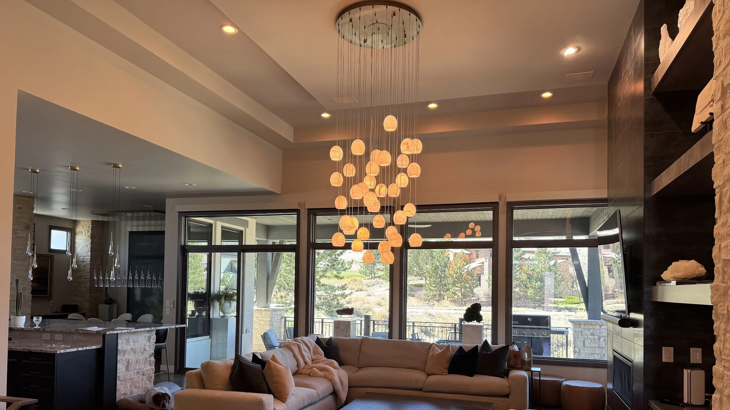Great room in a foothills home with a cluster chandelier of glowing globes, recessed lights and tall windows onto the pines