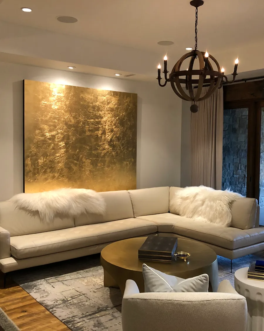 Iron chandelier and recessed lights over a cream sectional, with a gold-leaf painting lit on the wall